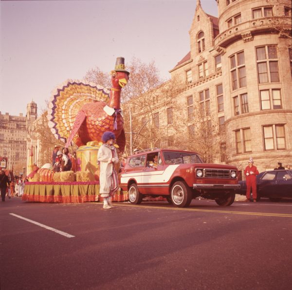 Thanksgiving Day Parade, either Macy's Parade in Manhattan, New York, or Gimbels' Parade in Philadelphia, Pennsylvania. A red and white International Harvester Scout 4x4 is towing a float with a large turkey wearing a white collar and Pilgrim style hat. Bales of straw, pumpkins, and children and adults can be seen on the float. A clown is walking alongside the Scout truck, and in the background is a building with a stone facade.