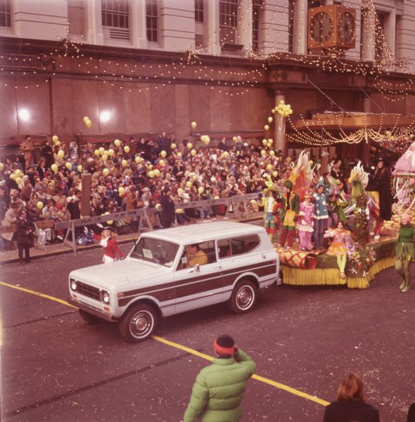 Macy's Thanksgiving Day Parade. The Macy's storefront can be seen in the background. A red and white International Harvester Scout 4x4 truck is towing a float. On the float a woman wearing a black cape and pointed black "witch cap" is seated on a multi-colored horse, with another horse and children in colorful costumes arranged around her. A woman in white pants, red jacket, dark blue tri-corner hat, and white gloves with blue trim is walking beside the Scout. Spectators with yellow balloons and uniformed police officers are watching from the sidewalk.