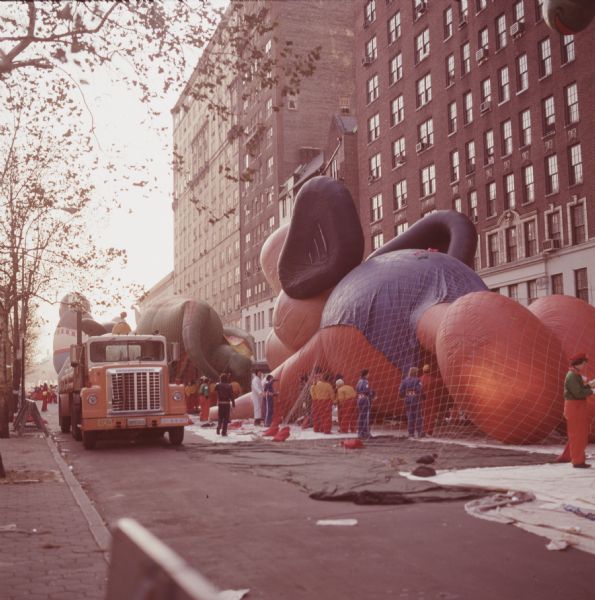 People EW working with floats for a Thanksgiving Day Parade, either Macy's Parade in Manhattan, New York, or Gimbels' Parade in Philadelphia, Pennsylvania. Workers are standing around nets holding down a balloon based on the cartoon character Mighty Mouse, and another character colored gray that may be Dumbo. A white and orange truck is parked in the street, and what appears to be apartment buildings are in the background. Trees and a sidewalk are on the right.