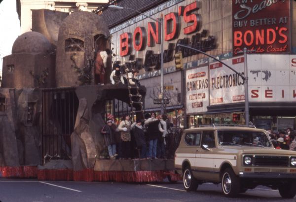Thanksgiving Day Parade, either Macy's Parade in Manhattan, New York, or Gimbels' Parade in Philadelphia, Pennsylvania. A tan and white International Harvester Scout 4x4 tows a parade float which resembles a cave-like structure with bars on two sides below and mud huts with domed tops above. Children on the lower level are gathered at the front of the float around a ladder, and children on top are dressed in what may be caveman costumes. A sign for Bond's department store is in the background.