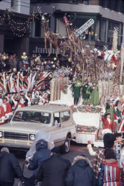 Macy's Thanksgiving Day Parade. A cream and white International Harvester Scout 4x4 is pulling a float with a Santa Claus theme. On the float, a group of reindeer are rising at an angle with Santa Claus in a sleigh behind. On the side of the float are children dressed in green as elves, and women in white clothes with red skirts and capes with white trim. Yellow balloons are rising into the air around the float. Alongside the float is a band in Scottish dress and spectators. Some of the band members are holding flags.