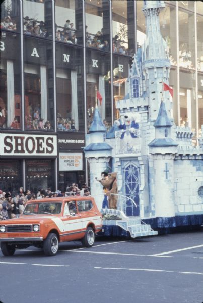 Thanksgiving Day Parade, either Macy's Parade in Manhattan, New York, or Gimbels' Parade in Philadelphia, Pennsylvania. An orange and white International Harvester Scout 4x4 is towing a Disney castle themed parade float. Goofy and Pluto are at the front of the float, while the Shaggy Dog, dressed in a blue jacket and tie, is on a balcony. Spectators are along the sidewalk in the background, and are also watching from large glass windows of a three-story storefront.