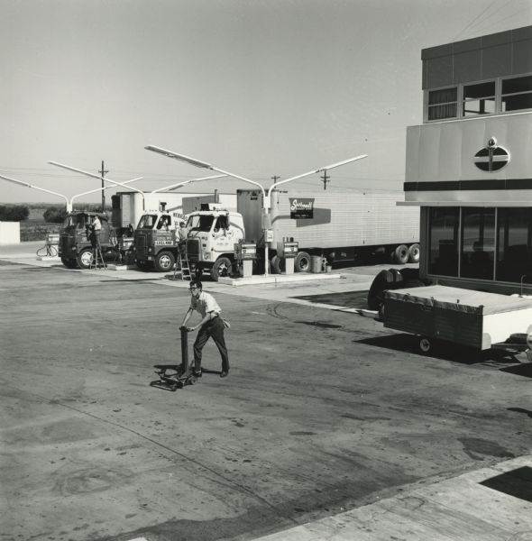 Mechanic pushing a hydraulic car jack at an American Oil Company truck stop. Three trucks are parked at the gas pumps in the background.