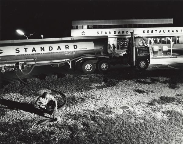 Elevated view at night of man filling an underground tank with a hose from a Standard Oil fueling truck. Behind the truck is an American Oil Company truck stop and restaurant.