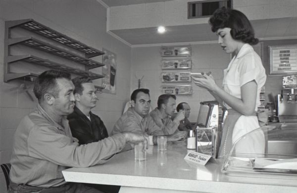 A waitress serves five male truck drivers seated along a diner counter at a truck stop. The original caption reads: "Service to a 'T' awaits drivers in a hurry at the special trucker counter. Stamping a red 'T' to orders gets the fastest service possible."