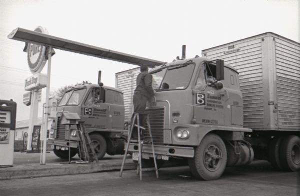 Gas attendant Sam Fry cleans the windshields of two International Harvester trucks parked near gas pumps. The original caption reads: "UPPER STORY. Gas Attendant Sam Fry truly gets a special lift out of cleaning the huge glass windshields of these twin Internationals."