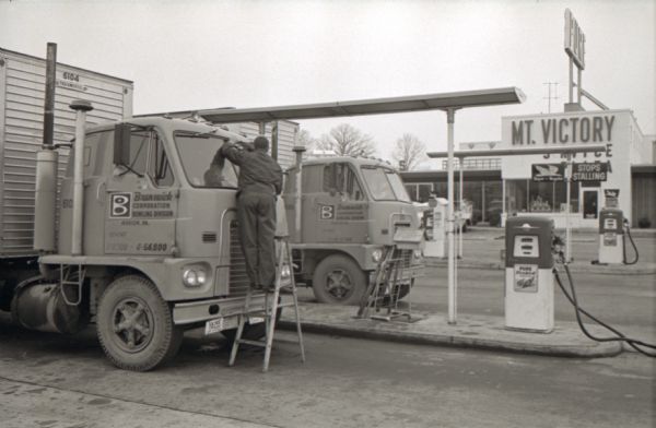 Gas attendant Sam Fry stands on a ladder to clean the windshields of an International Harvester truck parked near a gas pump at a truck stop. The original caption reads: "UPPER STORY. Gas Attendant Sam Fry truly gets a special lift out of cleaning the huge glass windshields of these twin Internationals."