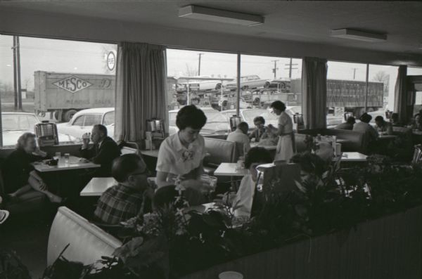 Waitresses serve tables of customers seated for a meal inside the Mt. Victory truck stop. The caption on a similar photograph reads: "Service to a 'T' awaits drivers in a hurry at the special trucker counter. Stamping a red 'T' to orders gets the fastest service possible."