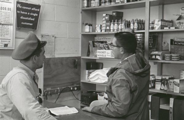 A worker helps a customer from across the counter at the general store in the Mt. Victory truck stop. A caption on a similar photograph reads: "Bigger than a football field, service plaza at Mt. Victory, Ohio, illustrates the upswing in accommodations provided for today's truck driver."