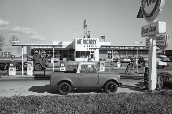 Exterior view of the Mt. Victory truck stop. An International Scout is parked in the foreground. Fuel pumps, a truck trailer, and the front of the plaza, including windows of the restaurant, are in the background.