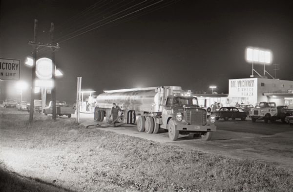 View at night of man filling an underground tank with a hose from a  fueling truck at the Mt. Victory truck stop. The caption on a similar photograph reads: "Most important, there's security in knowing it's always open. Travelcase in hand, Roger heads for a hot meal and relaxation."