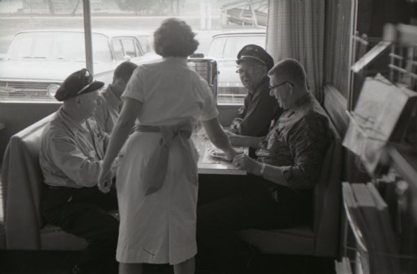 A waitress serves four truck drivers sitting in a booth in the Mt. Victory truck stop dining room. Against the window of the booth is a tabletop jukebox.