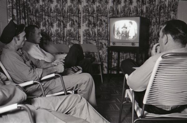 A group of truck drivers watch television while relaxing at the Mt. Victory truck stop. The caption on a similar photograph reads, "Bigger than a football field, service plaza at Mt. Victory, Ohio, illustrates the upswing in accommodations provided for today's truck drivers."