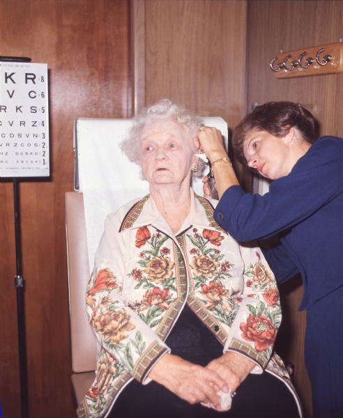 A nurse IS examinING an elderly woman in a mobile health unit operated by the Alfred University School of Nursing. The mobile health unit traveled around communities in Allegany County, New York providing health care services and education. The elderly woman is wearing a top with vivid flowers, and the nurse is wearing dark blue clothing. Behind them are an eye chart and hooks to hang coats on. The photo caption reads: "Examination Mrs. Joan Cameron, R.N., was giving Mrs. Ruby Henry a sampling of professional services dispensed by vehicle's staff."