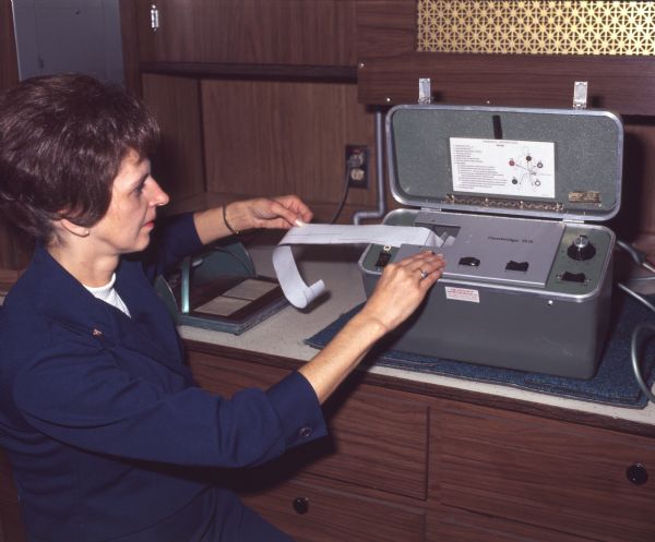 Dr. Virginia Barker, Nursing School Dean at Alfred University, is reading the results of an electrocardiograph in a mobile health unit.  The mobile health unit travelled around communities in Allegany County, New York, providing health care services and education. Dr. Barker is wearing a dark blue uniform, and the electrocardiograph is on a cabinet of drawers. There is a gold-colored screen on the wall behind the machine.