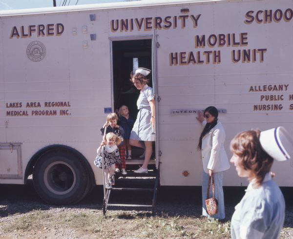 Children are standing on the steps of the Alfred University School of Nursing Mobile Health Unit. The mobile health unit traveled around communities in Allegany County, New York, providing health care services and education. Two nurses in uniform and a third woman with a purse are standing outside the vehicle, which was built on an International Harvester model 1853-FC chassis.