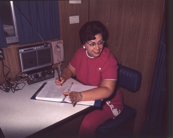 A woman is sitting at a table filling out a ledger. She is wearing red clothing with white piping, and is wearing eyeglasses. She is probably a nurse or nursing student working at the mobile health unit operated by the Alfred University School of Nursing. On the table is an AM/FM radio, and mounted on the wall are a tape recorder and phone.