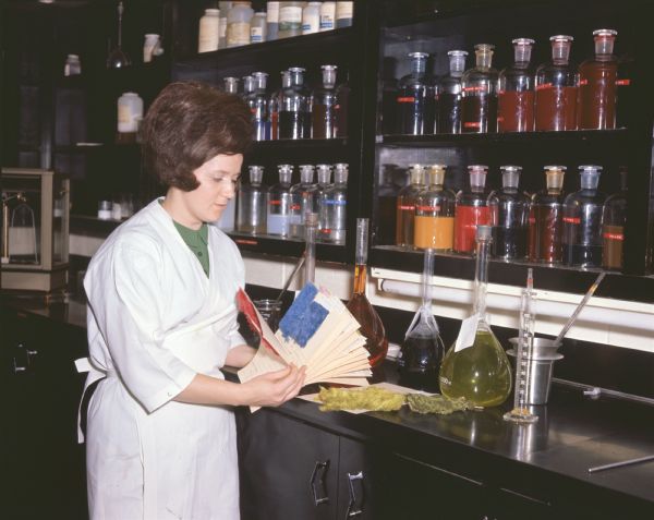 A female technician is at a workbench testing dyes for nylon carpet. The woman is wearing a white lab coat and apron, and has a beehive or bouffant hairstyle. She is holding paperwork that have small swatches of carpet attached. Several flasks with variously colored liquids in them are on the bench, and glass jars with variously colored chemicals are on shelves on the wall. Photo captions reads: "Because of nylon's versatility, some carpet lines come in as many as 30 colors. Here technician tests colors for dyes at Delta Finishers, Inc."