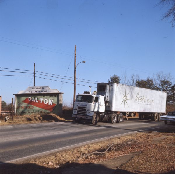Color photo of an International Harvester truck model C-OF4070A parked by a sign welcoming visitors to Dalton, Georgia. The sign reads: "Population Met. Area 28,000 Welcome to Dalton, Tufted Textile Center of the World Dalton Chamber of Commerce." The truck is operated by Loomtex Mills, Inc., whose logo can be seen on the side of the trailer. There is dry grass and various brown foliage around the sign and along the side of the road. Photo caption reads: "Steady stream of International trucks rolls past sugb trumpeting Dalton's global affluence. Pictured C-OF4070A is fielded by Loomtex Mills, Inc."
