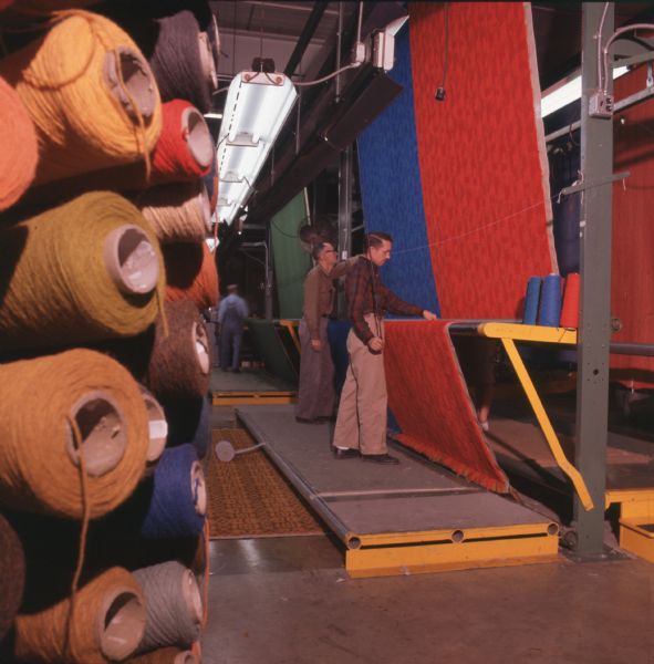 Workers in a carpet factory apply latex backing to hold yarn in place. The workers are standing on a slightly raised platorm, and the carpet is being fed vertically, horizontally across rollers, and then vertically again. Large spools of yarn in a variety of colors can be seen, and large industrial lights hang from the ceiling. Photo caption reads: "Dalton carpetmaking plants abound with sights like this. In finishing operation, latex applied to backing locks dense pile of yarn into place. (Monsanto Company Photo)"