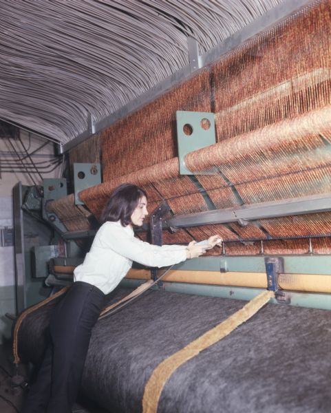 A woman is working on a tufting carpet loom. She is leaning into the loom which has a large number of individual strands of yarn on top, and gray material, which may be backing, on the bottom. The woman is holding a gray, box-like control button in her hand, possibly to operate the loom.  
