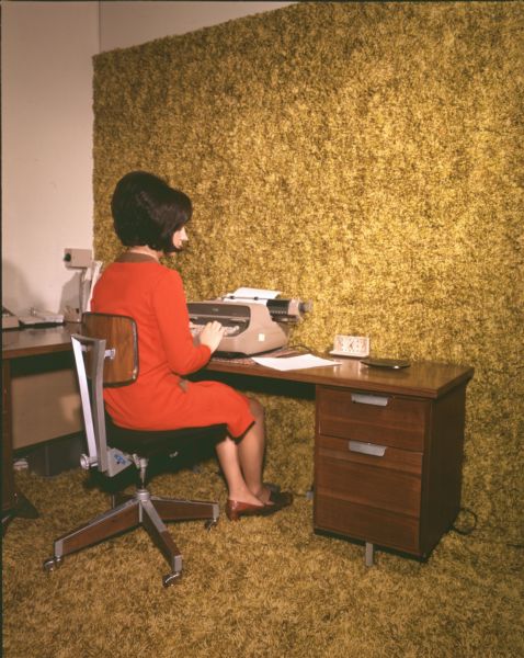 A woman wearing a red dress, brown shoes, and  a beehive or bouffant hairstyle is sitting on a rolling chair with an adjustable back at a desk. She is composing a letter on a typewriter. On the he desk is a clock and a small, shallow dish, possibly an ashtray. The floor and part of the wall around the desk are covered in a brownish-yellow-green colored carpet.