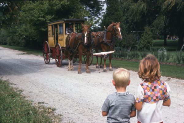 Two children are standing at the side of a gravel road looking at a man driving a horse-drawn enclosed carriage. The carriage had been used to transport children to and from school in 1915. The carriage was brought out by Kickert School Bus Line, Inc. on its 60th anniversary. Kickert originally serviced the Southern Cook county area.