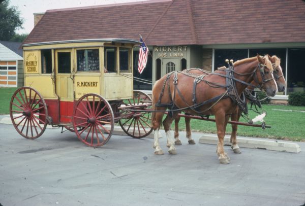 A horse-drawn enclosed carriage painted yellow, with the undercarriage and wheels painted red, is parked outdoors in front of a building. On the side of the carriage are painted signs reading: "Kickert Bus Lines," "Service to McKinley School" and "R. Kickert VEH. No. 1." A flag is mounted on the building beside the door which has a sign that reads: "Kickert Bus Lines."