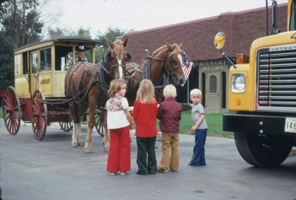 Four small children are standing in a parking lot looking at a horse-drawn enclosed carriage. The carriage had been originally used to transport children to and from school in 1915. An older man is sitting at the front of the carriage, which has "Service to McKinley School" and "R. Kickert VEH. No. 1" written on the side. The front of a school bus with an International Harvester Loadstar chassis is on the right. In the background is a building with a "Kickert Bus Lines" sign above the door.