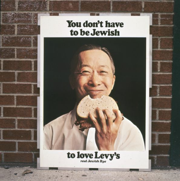 A sign shows a smiling Asian American man holding a rye bread sandwich in an advertisemnt for Levy and Sons Bakery of New York, New York. The sign reads: "You don't have to be Jewish to love Levy's real Jewish Rye." The ad was featured on the side of delivery trucks, subway and railway platforms, on television, and in newspapers and trade books. The photo caption reads: "Plainly evident in sampling at top, form plays the sandwich-eater illustrations with unaltered candor. Ad campaign's effect on consumers. Rye loaf's one of the most popular in the East."
