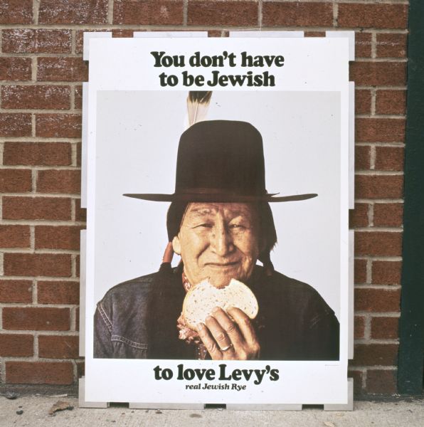 A sign shows a smiling Native American man holding a rye bread sandwich in an advertisment for Levy and Sons Bakery of New York, New York. The man is wearing a denim jacket, has long hair in two braids, and a black hat with a feather. The sign reads: "You don't have to be Jewish to love Levy's real Jewish Rye." The ad was featured on the side of delivery trucks, subway and railway platforms, on television, and in newspapers and trade books.