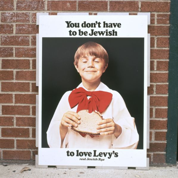 A sign shows a smiling choir boy holding a rye bread sandwich in an advertisment for Levy and Sons Bakery of New York, New York. The boy is wearing an altar boy's vestment with a large red bow. The sign reads: "You don't have to be Jewish to love Levy's real Jewish Rye." The ad was featured on the side of delivery trucks, subway and railway platforms, on television, and in newspapers and trade books. The photo caption reads: "Plainly evident in sampling at top, form plays the sandwich-eater illustrations with unaltered candor. Ad campaign's effect on consumers. Rye loaf's one of the most popular in the East."
