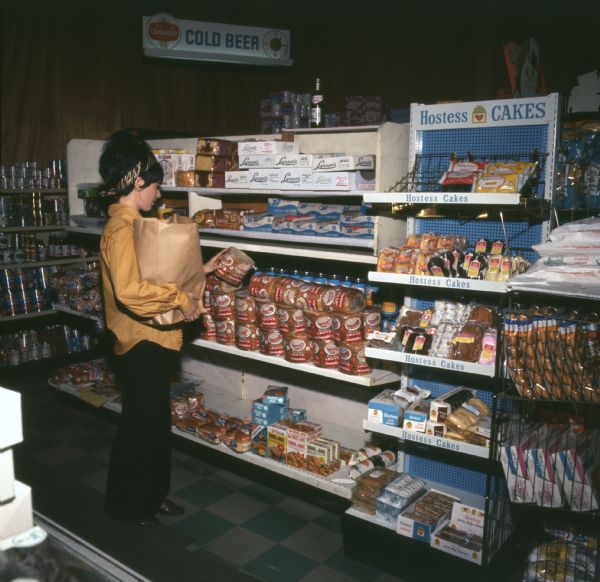 Woman standing in a store aisle examining a loaf of Levy and Sons Bakery rye bread. She is wearing a yellow shirt, colorful headband, and a beehive or bouffant hairstyle. A variety of products can be seen, including Shake 'N Bake and a wide range of Hostess snack cakes. A sign reading "COLD BEER" is in the background. The photo caption reads: "Plainly evident in sampling at top, form plays the sandwich-eater illustrations with unaltered candor. Ad campaign's effect on consumers. Rye loaf's one of the most popular in the East."