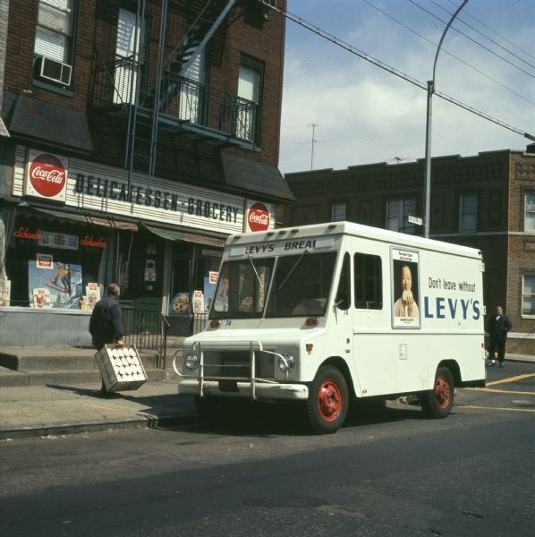 View from street of a man delivering Levy and Sons Bakery rye bread to a New York City delicatessen and grocery store. The truck is an International Harvester model MA-1500 Metro, and has "Levy's Bread" written on the front. On the side of the truck is written: "Don't Leave Without Levy's" and there is also a framed advertising poster showing a smiling Chinese man eating a rye bread sandwich. Signs in the storefront depict a skier jumping and say: "Enjoy Coca-Cola," "Schaefer," "Cold Beer Here." On the corner is a street sign for Onderdonk Avenue. The photo caption reads: "Baker's use of Metro truck walls for display of distinctive posters is exemplified as camera catches route salesman servicing a stop in Ridgewood, Queens, N.Y. Uniformly marked, fleet handling Levy's store-to-store deliveries includes 36 new MA-1500 International trucks."