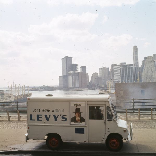 Elevated view of an International Harvester model MA-1500 Metro delivery truck is parked on a bridge overlooking a body of water and a city skyline, likely somewhere in New York City. On the side of the truck is written: "Don't Leave Without Levy's." There is also a framed  advertising poster showing a smiling Native American man. The advertising poster reads: "You don't have to be Jewish to love Levy's real Jewish Rye." In the background are two ships with cranes on their decks, one with [?]inea Fassio written on the side. In the far background are skyscrapers.
