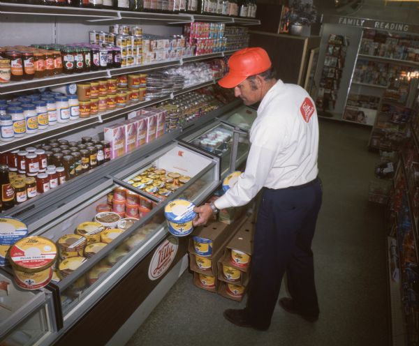 A routeman for Lilly Ice Cream of Bryan, Texas, is stocking a freezer cabinet with a sign that reads: "Lilly Ice Cream." The man is wearing dark blue slacks, a white shirt with a red patch saying "Lilly Dairy Products" on the back, and a red trucker cap. The shelf above the freezer cabinet shows a wide variety of grocery products, including Miracle Whip, Peter Pan Peanut Butter, Welch's Jam and Jelly, Planter's Peanuts, Nabisco Comet Cups, and Jell-O. A book rack and assorted magazines with a sign that reads: "Family Reading" is in the background. The photo caption reads: "Stocking of store freezer cabinets with Lilly product is a routine strongly supported by firm's Loadstar trucks. Routemen work from 1600's wide side-loading bodies."