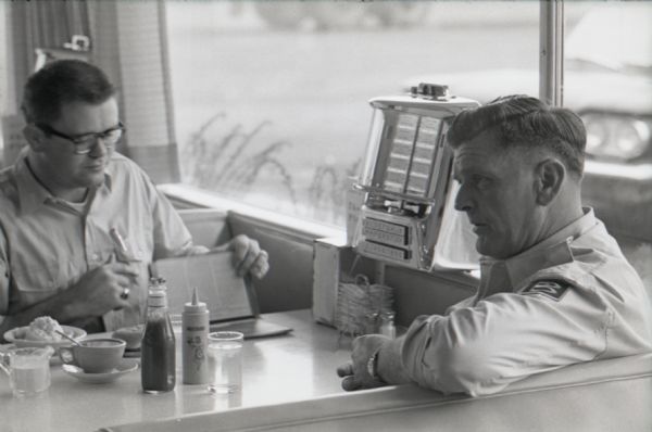 Two truck drivers sit on either side of a booth at the Mt. Victory truck stop, or service plaza, while waiting for a meal. At the end of the booth near the window is a tabletop jukebox.