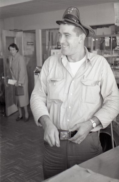 A truck driver stands in the Mt. Victory truck stop. He wears a hat, wristwatch, and a decorative metal belt buckle. A woman stands in a doorway on the left in the background.