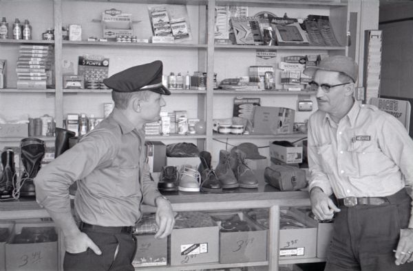 Two men converse in the general store area of the Mt. Victory truck stop. The man on the right is holding a cigarette. Shoes are arranged on the countertop, and various supplies are on display on shelving in the background.