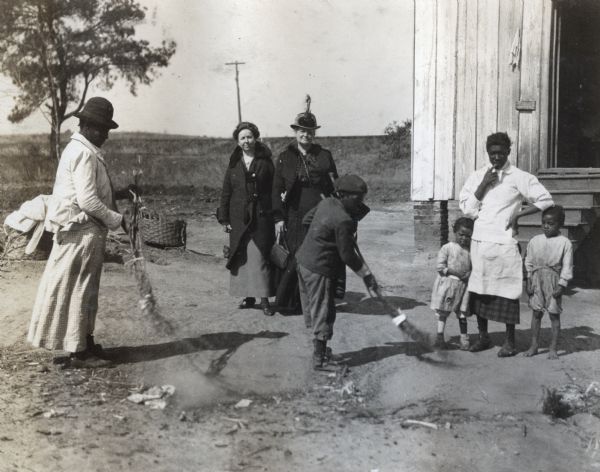 A woman and young boy sweep yard with "brush broom" while another woman with two children looks on. Two additional women, possibly Agricultural Extension employees watch as well. Original caption reads, "the brush broom made by tying a bundle of brush together, is used a great deal in the South, as it costs nothing to make and serves well as a broom for rough sweeping around the yard. A custom which most of the Southern people follow."