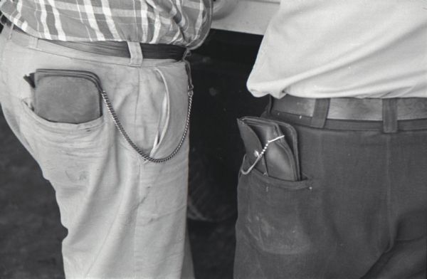 Close-up view of the back pockets of two men which hold their wallets that are chained to their belt loops. The men were truckers taking a break at the Mt. Victory service plaza. The caption on the photograph reads, "Precaution on the road carries over to personal safety. Truckers chain wallets to belts to guard against robbery while they're sleeping in truck cabs."