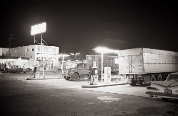Trucks line up at the gas pumps outside the Mt. Victory service plaza at night. The caption on a similar photograph reads, "Bigger than a football field, service plaza at Mt. Victory, Ohio, illustrates the upswing in accommodations provided for today's truck drivers."