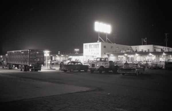 Trucks and automobiles parked outside the Mt. Victory service plaza at night. The caption on a similar photograph reads, "Bigger than a football field, service plaza at Mt. Victory, Ohio, illustrates the upswing in accommodations provided for today's truck drivers."