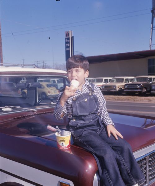 A five year old boy is sitting on the hood of a truck eating an ice cream cone. The boy is Joel Keith Strickland, and he is wearing denim bib overalls and a blue and white checkered long sleeve shirt. There is a pint of Lilly Ice Cream (likely vanilla) with an ice cream scoop in it beside him on the truck. An International Harvester dealership is in the background, and several vehicles can be seen parked along the side of the building. A yellow Volkswagen Bug is parked on the street.