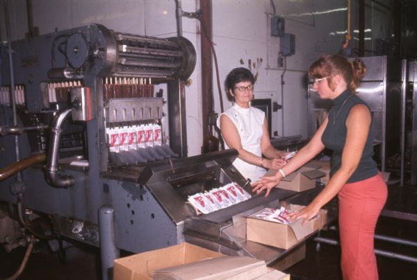 Two women are standing at the end of a machine which is packaging Lilly Ice Cream sticks. The machine is placing ice cream sticks in individual wrappers, and then they travel to the workers who are packing them into boxes. The women are wearing glasses and sleeveless shirts. One woman has her hair tied back into a bun. Other machinery is in the background.