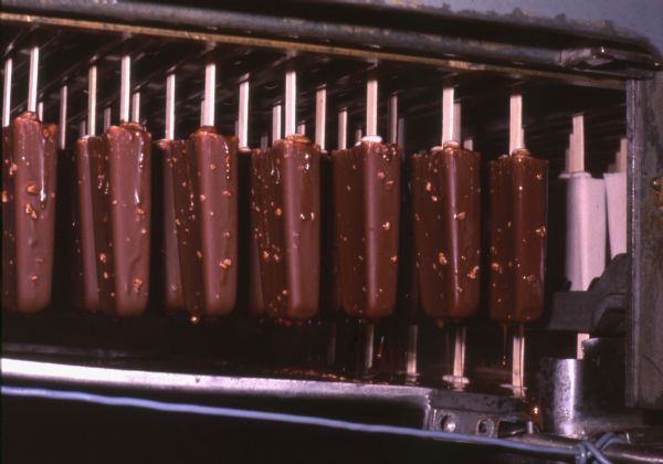 Ice cream sticks travel down a conveyor in the Lilly Ice Cream factory. Some of the ice cream sticks have been dipped in chocolate, and some have not. Chocolate can be seen dripping off some of the dipped ice cream sticks.