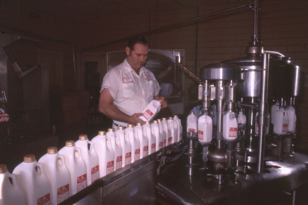 A man is standing near a machine filling milk bottles. A conveyor line in front of him is bringing empty milk bottles to the machine where they are filled and then taken away by another conveyor. The man is holding a bottle of milk and appears to be checking the lid. He is wearing a white shirt with two breast pockets, black belt, and white pants. The scene is likely from the Jersey Land dairy production plant.