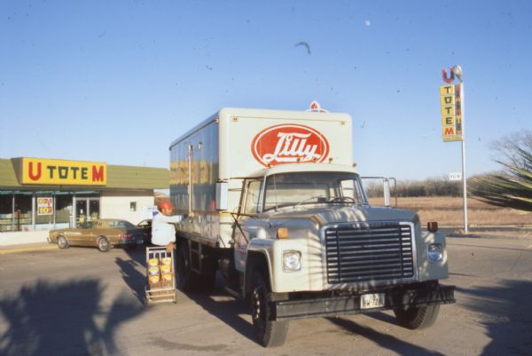 An International Harvester model 1600 Loadstar is parked at a U Tote M Food Store. The truck is a Lilly Ice Cream delivery vehicle, and a worker is unloading ice cream from the side door. A "U Tote M" sign with a stylized totem pole is on the right. Cars are parked in front of the storefront.