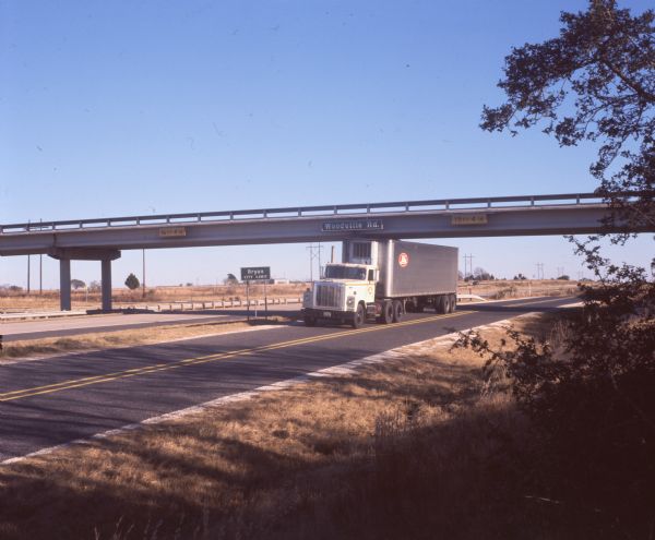 View from side of road of an International Harvester model F-4370 truck traveling down a Texas highway. Behind the truck is an overpass with a sign reading: "Woodville Rd." and "15 FT 4 IN." The truck is a delivery vehicle for Lilly Ice Cream of Bryan, Texas. A sign saying: "Bryan City Limit" is in the median.