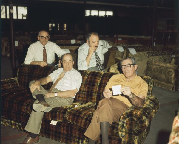 Four men, all brothers, posing in a warehouse full of sofas at Mastercraft Furniture Corporation. Two of the men are sitting on a sofa or couch, while two men behind them are leaning against the back. Three of the men are wearing dress shirts and ties, two are wearing glasses, and one is looking at a piece of paper. Left to right: Sam, David, Maury and Julius Katzman.